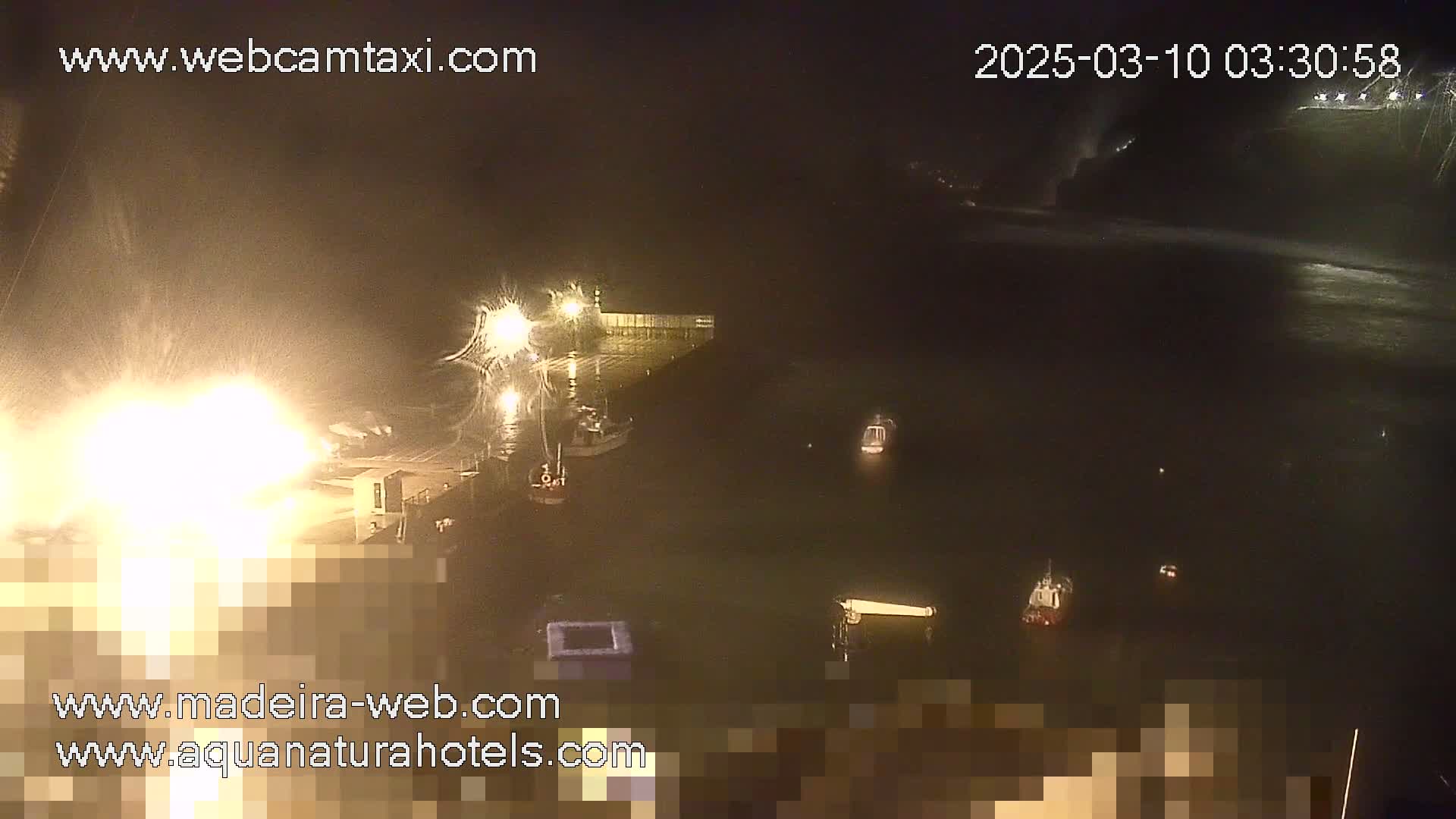 A nighttime view of a harbor with several boats moored, illuminated by bright lights, under cloudy conditions.