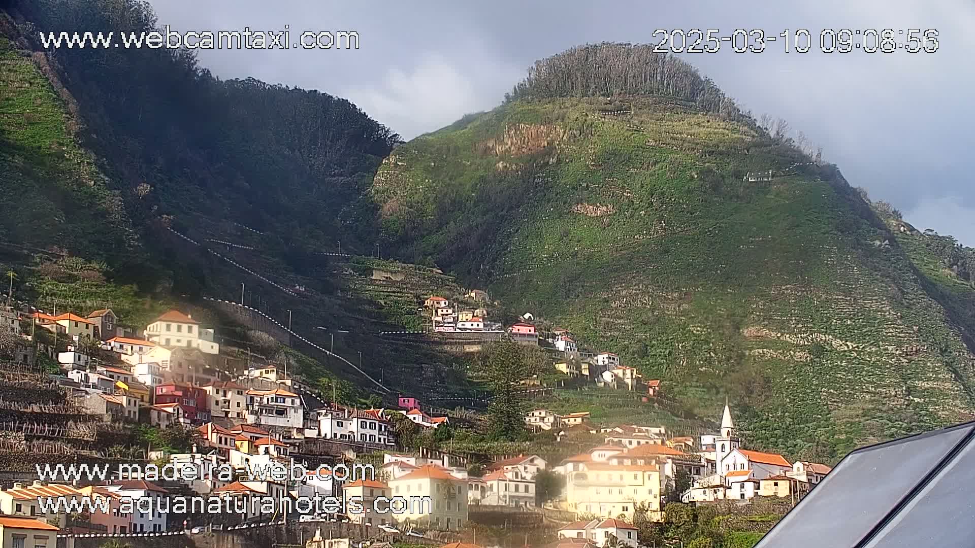 A village of low, colorful buildings clings to the side of a steep, green hill under partly cloudy skies.