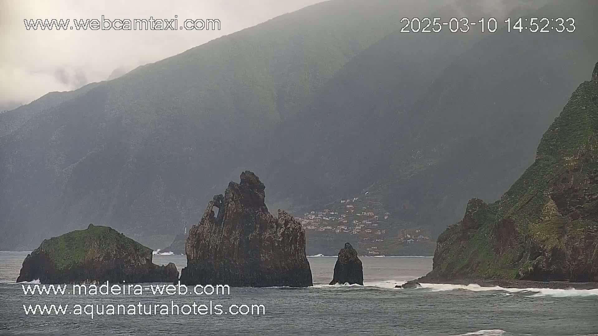 A misty, overcast day reveals a rocky coastline with several prominent rock formations jutting from the ocean near a small village nestled against steep, green mountains.