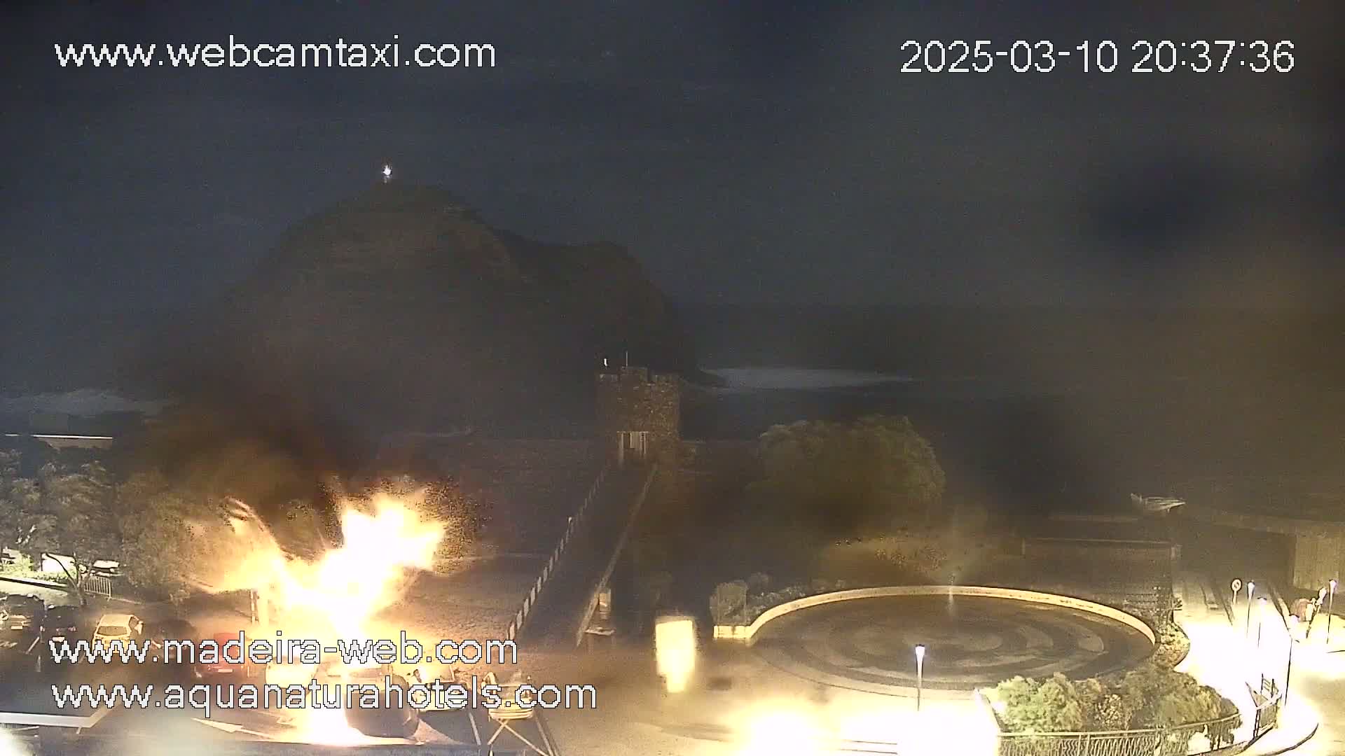 A nighttime coastal scene shows a large fire burning near a stone structure and a circular plaza, with a dark, calm ocean and a hilltop in the background.