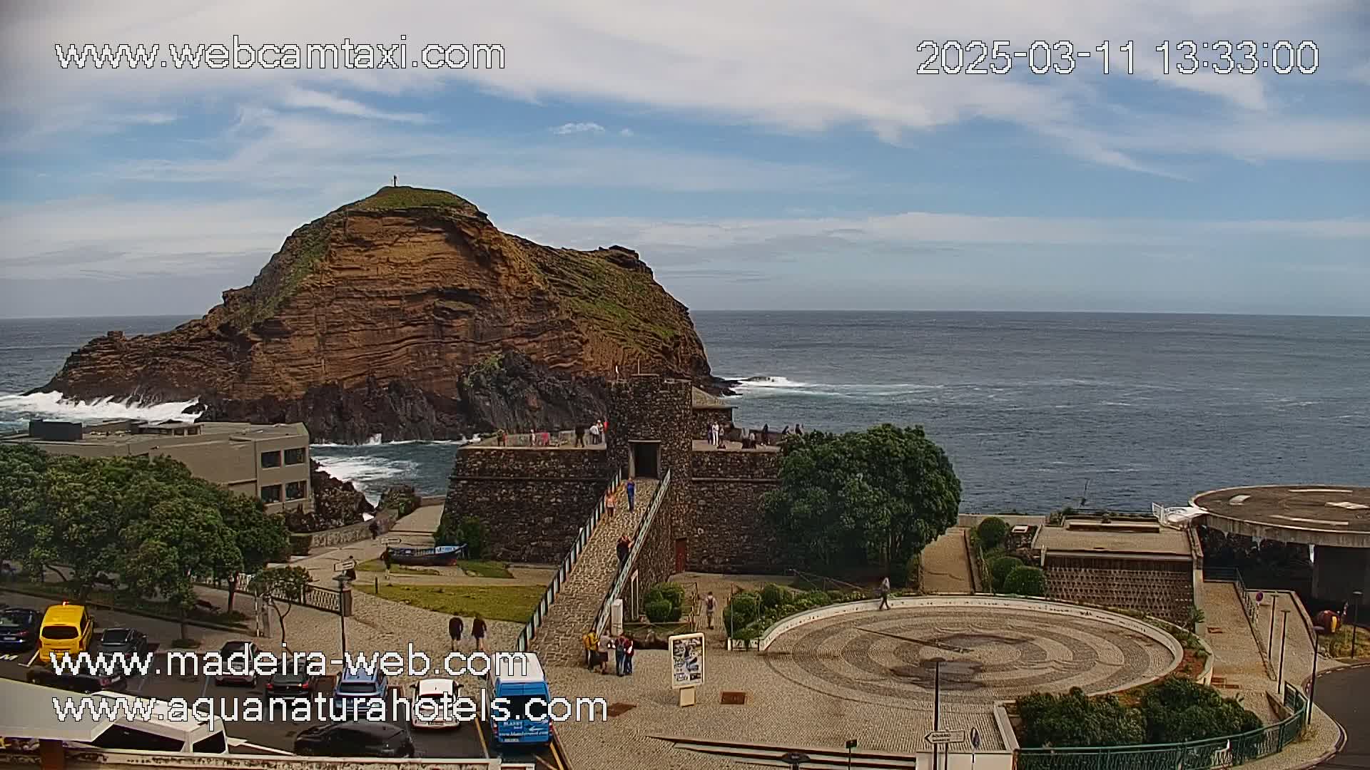 A rocky coastal headland with a stone structure and surrounding plaza is visible under partly cloudy skies, with a calm ocean in the background.