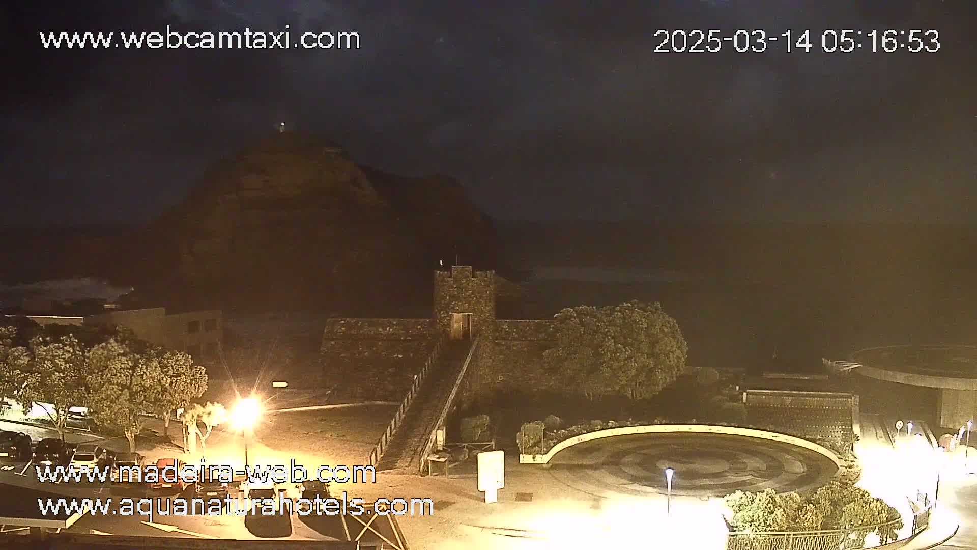 A nighttime, coastal view shows a stone structure near a circular plaza, with a hill and ocean in the background under a dark sky.