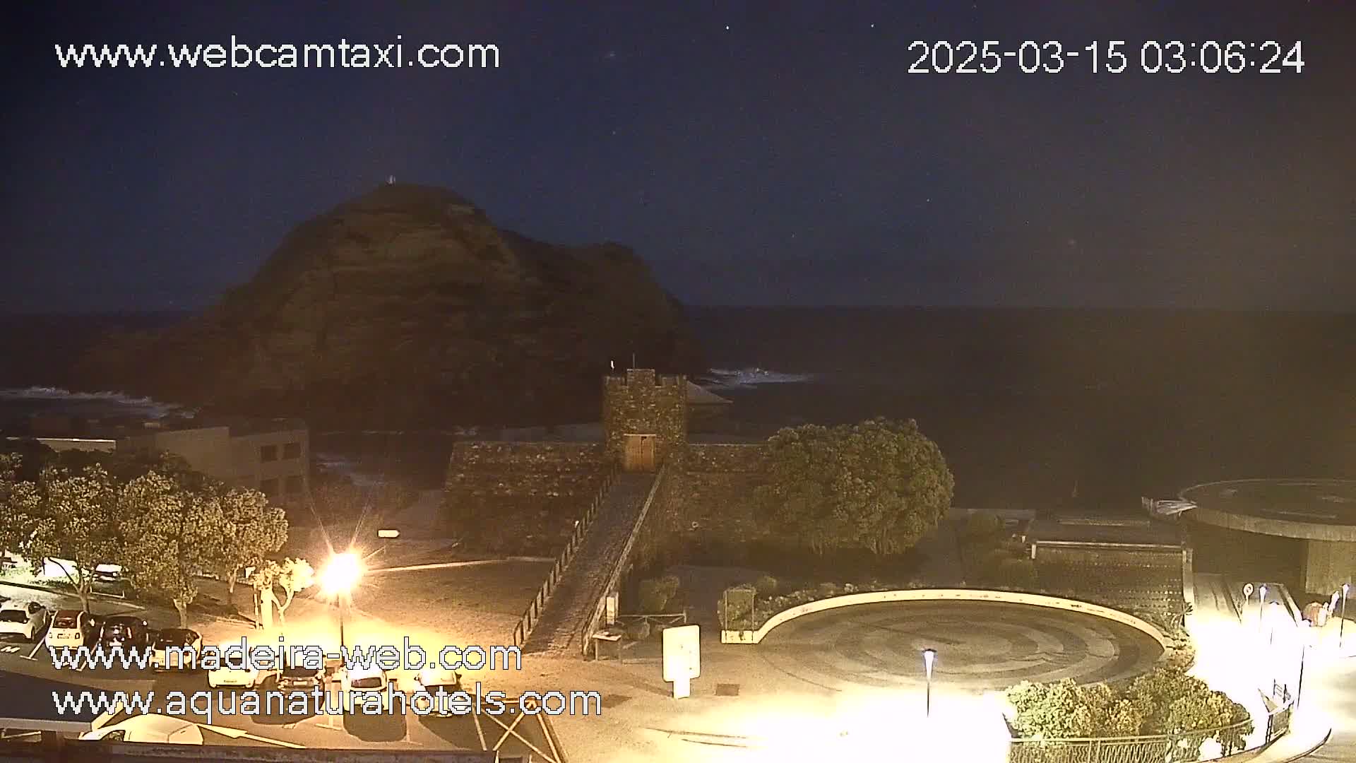 A nighttime coastal scene shows a dark, rocky hill, a stone structure resembling a small castle, a paved area with parked cars, and calm ocean waves under a clear, starry sky.