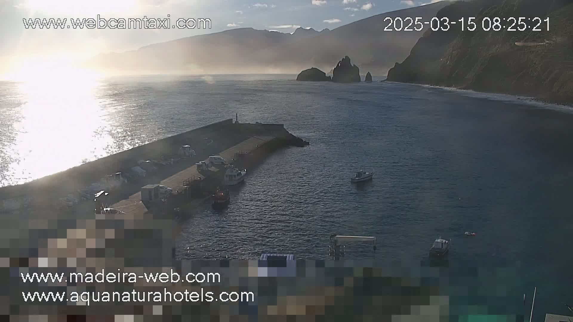 A hazy sunrise illuminates a calm harbor with several small boats moored near a rocky coastline and distant mountains.