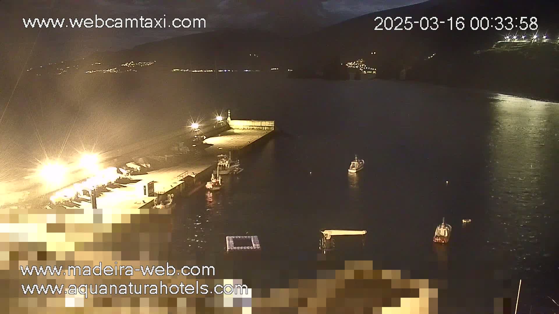 A nighttime view of a harbor with several small boats moored near a brightly lit dock, under a dark sky.
