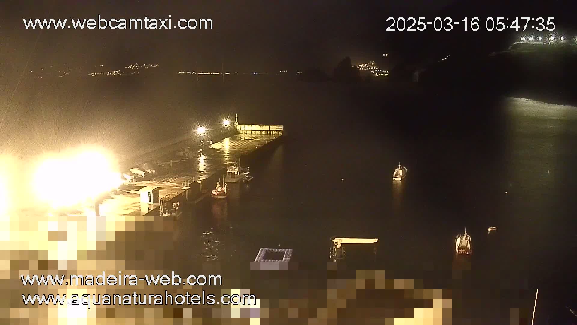 A nighttime harbor scene with several boats moored near a brightly lit dock, under somewhat stormy conditions.