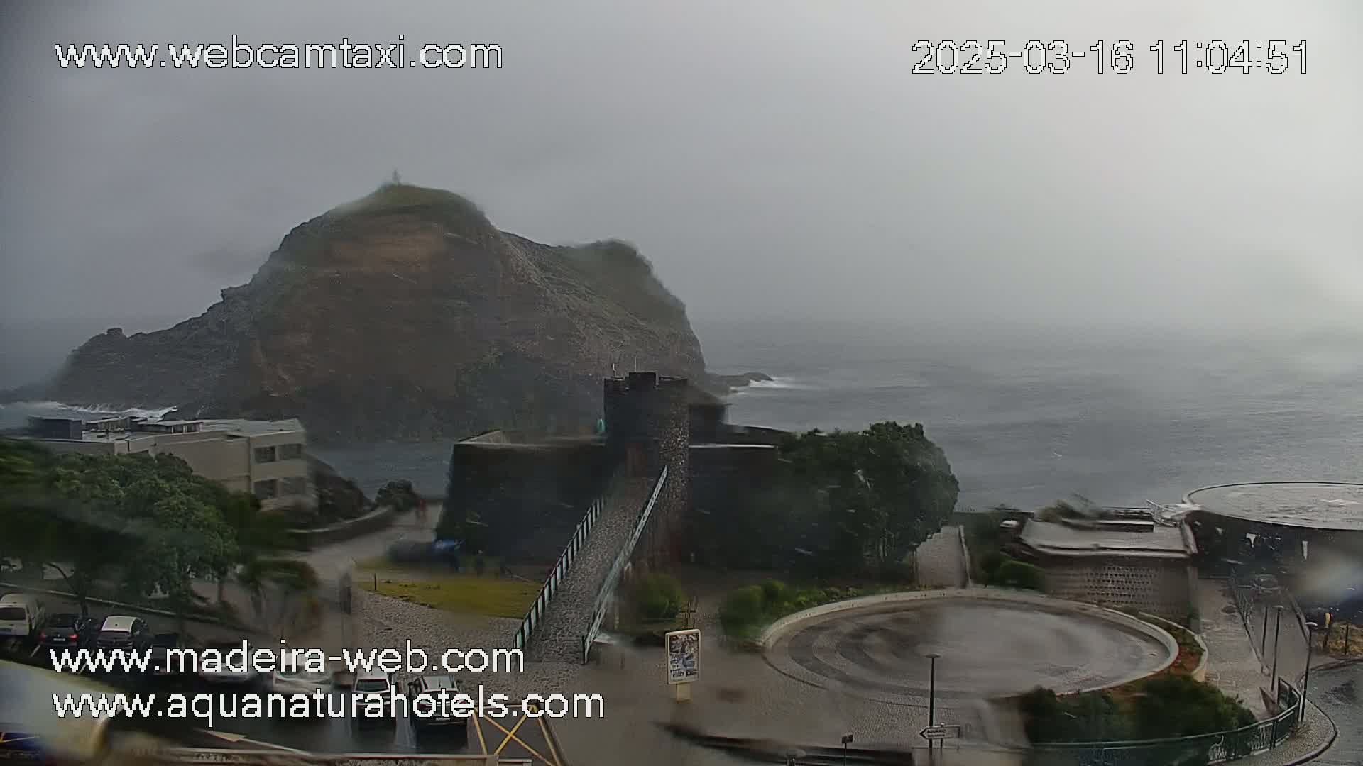 A rain-swept coastal scene shows a rocky outcrop with a building and bridge, overlooking a turbulent ocean.