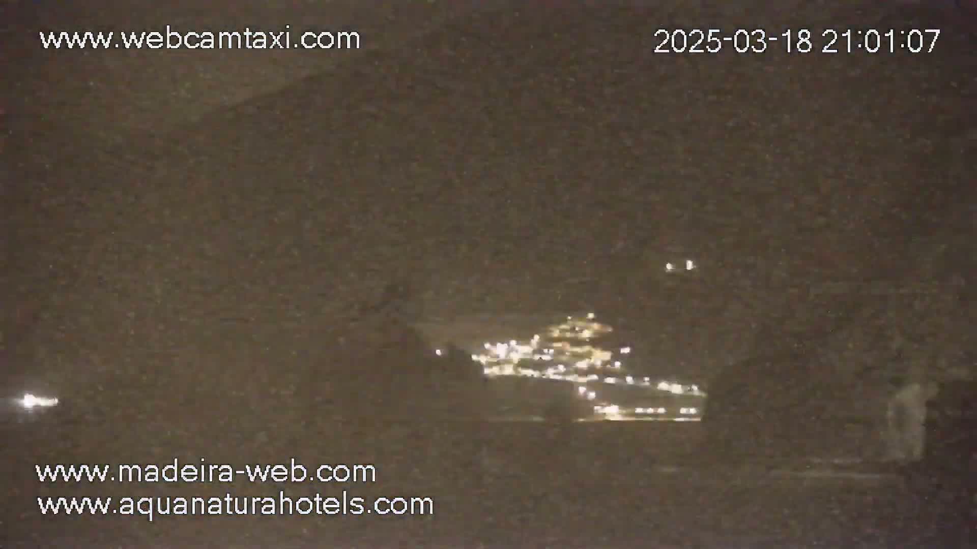 A nighttime long shot shows a dimly lit town or city across a body of water under a dark sky.