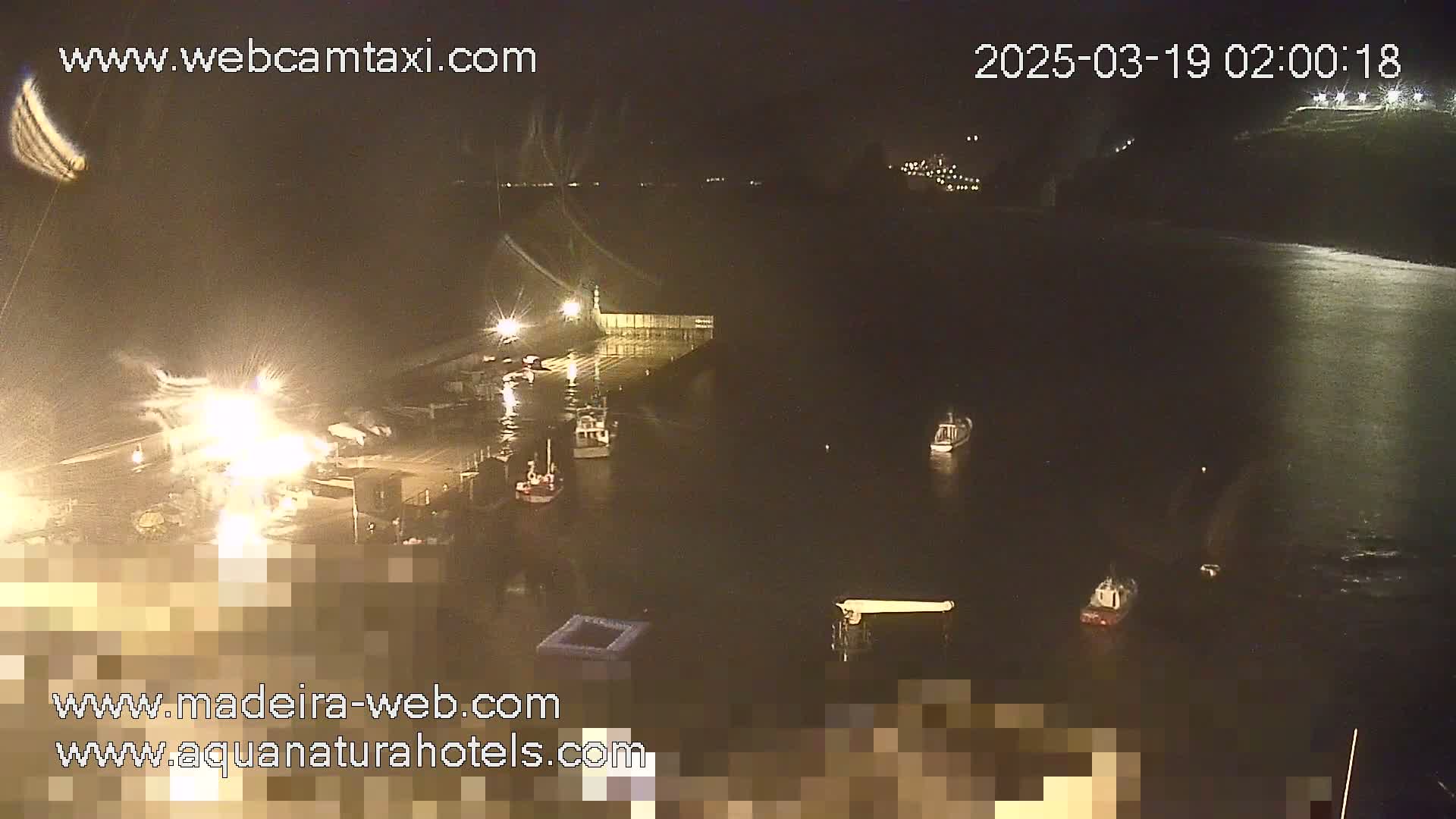 A nighttime long shot of a harbor with several boats moored, illuminated by artificial lights under partly cloudy skies.