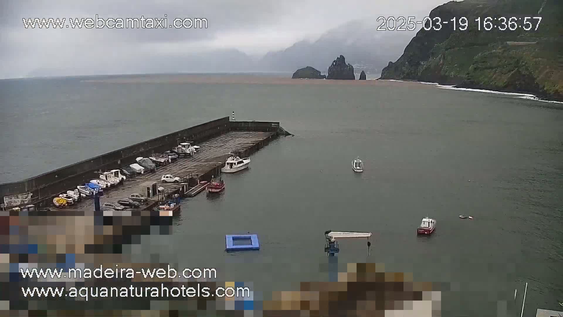 A small harbor with several boats docked, under an overcast sky, shows a rocky coastline in the distance.