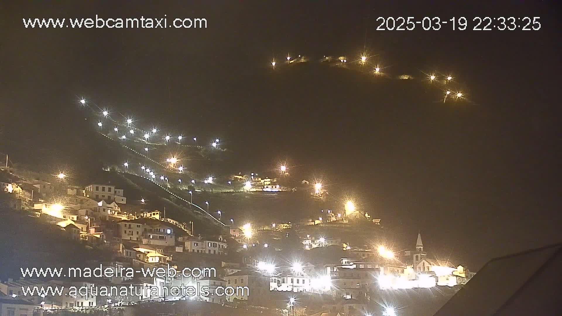 A hillside village is illuminated at night by numerous streetlights, appearing clear and calm under a dark sky.