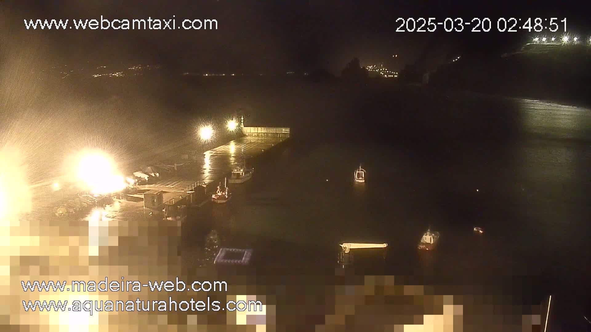 A nighttime view of a harbor with several boats, a dock, and a bright light spraying water, under rainy conditions.