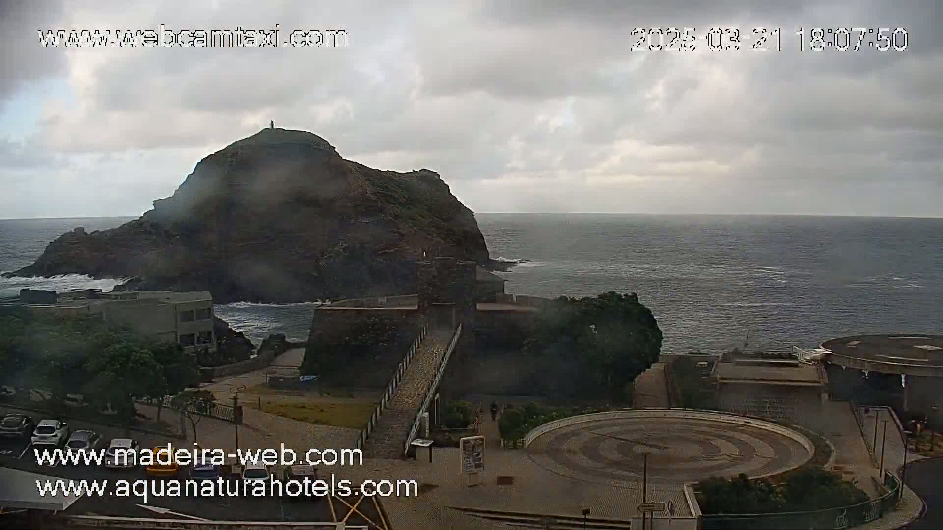 A coastal scene under an overcast sky shows a dark, rocky hill jutting into a choppy ocean, with a stone structure and paved area below.