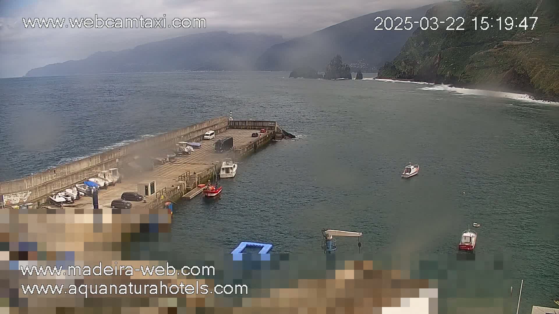 A small harbor with several boats and cars is nestled against a rocky coastline under an overcast sky.
