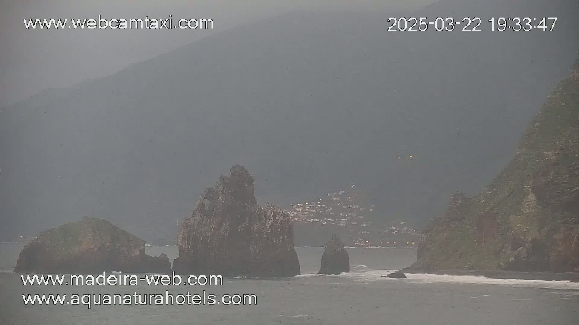 A hazy, overcast view shows rocky outcrops in the ocean near a distant town nestled against a mountainside.