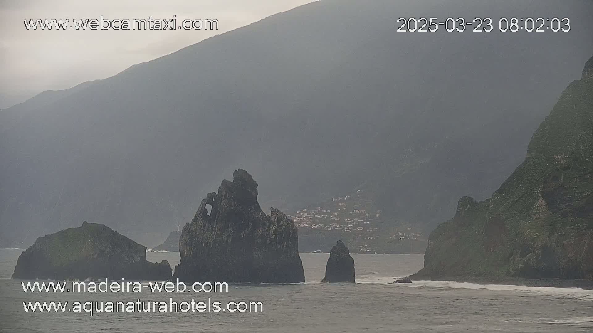 Dark, rocky coastal formations jut out from a hazy, overcast ocean near a village nestled against a mountainside.