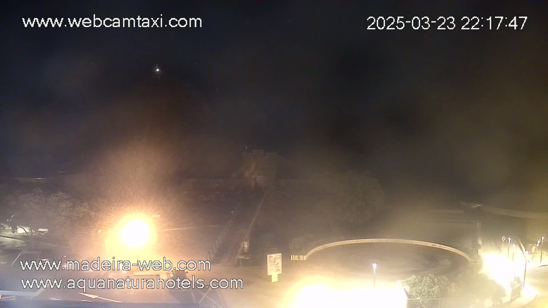 A nighttime, hazy view of a roadway and a circular area, possibly a roundabout, with some vegetation and bright lights visible.