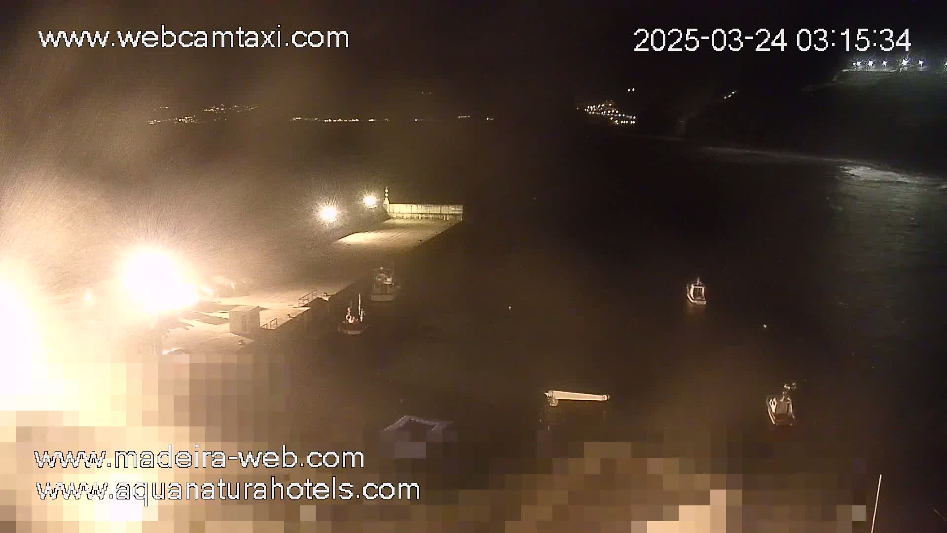 A nighttime view of a harbor with several boats moored at a pier, under heavy, blowing snow or spray.