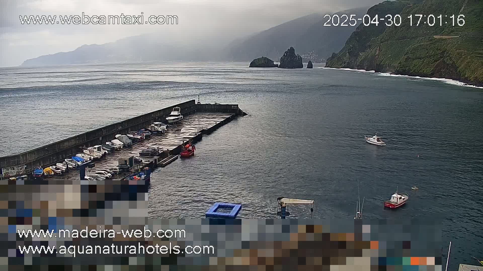 A harbor with numerous boats docked, a few boats in the water, and a mountainous coastline in the distance under an overcast sky.