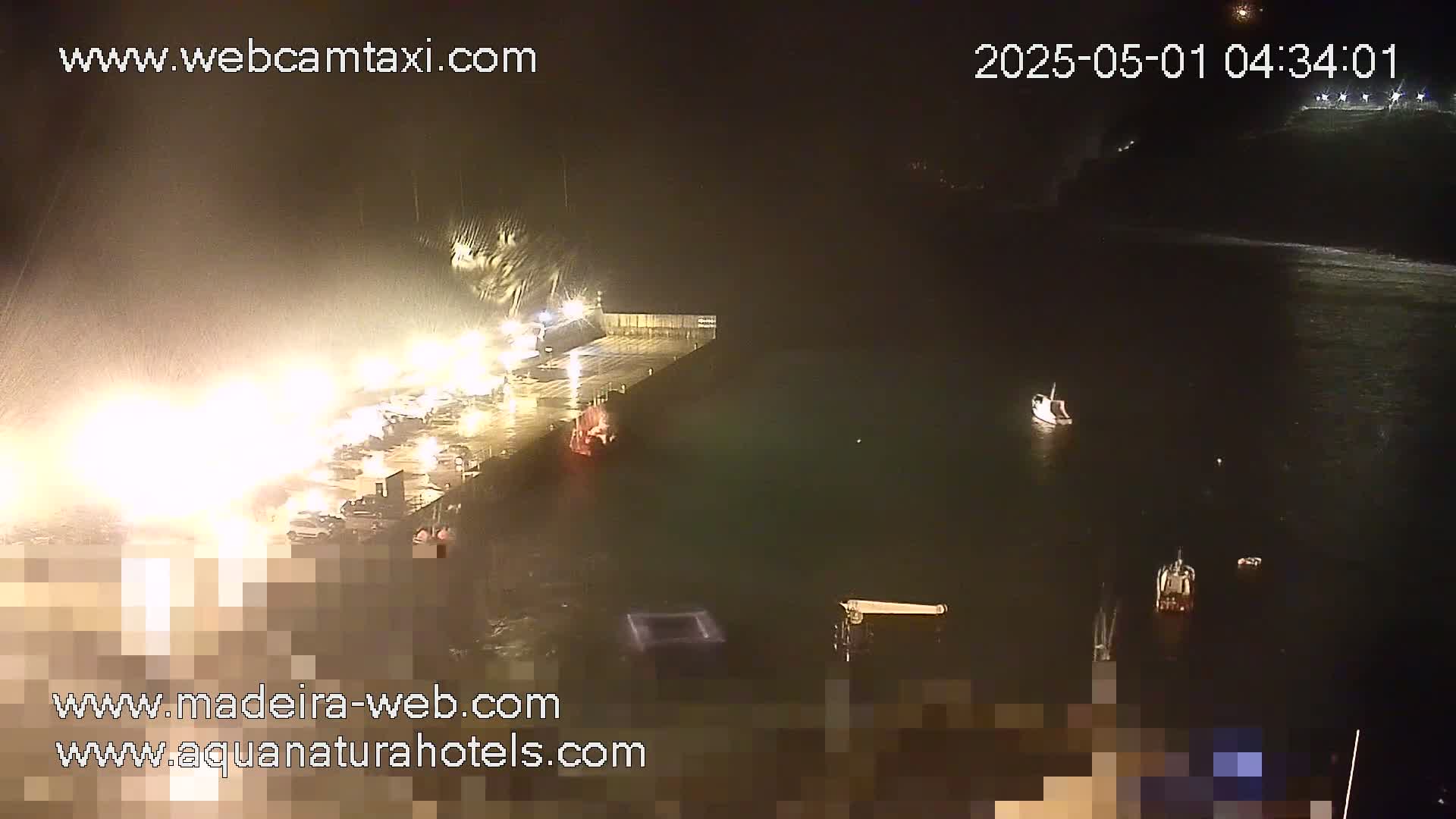 A nighttime, rainy scene shows a harbor with several boats and a dock illuminated by bright lights.