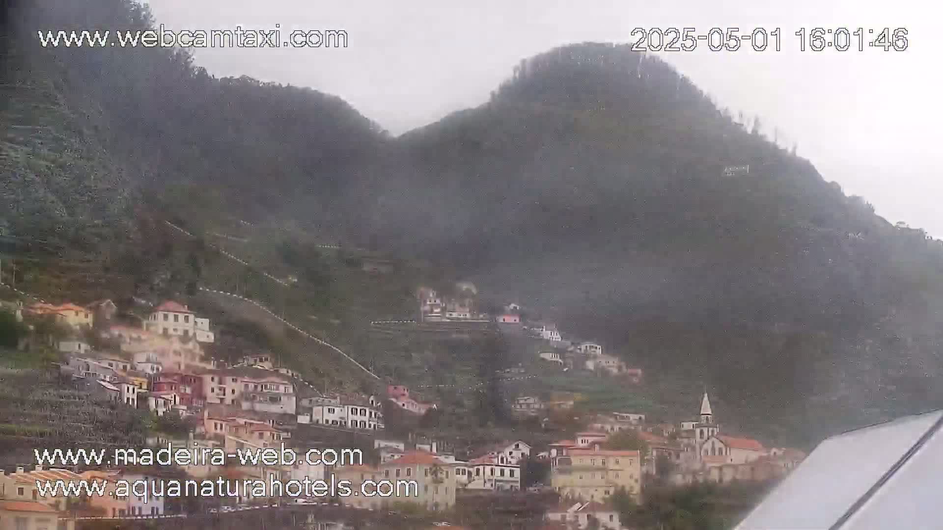 A village nestled on a hillside is shrouded in mist and light rain, with mountains rising behind it.