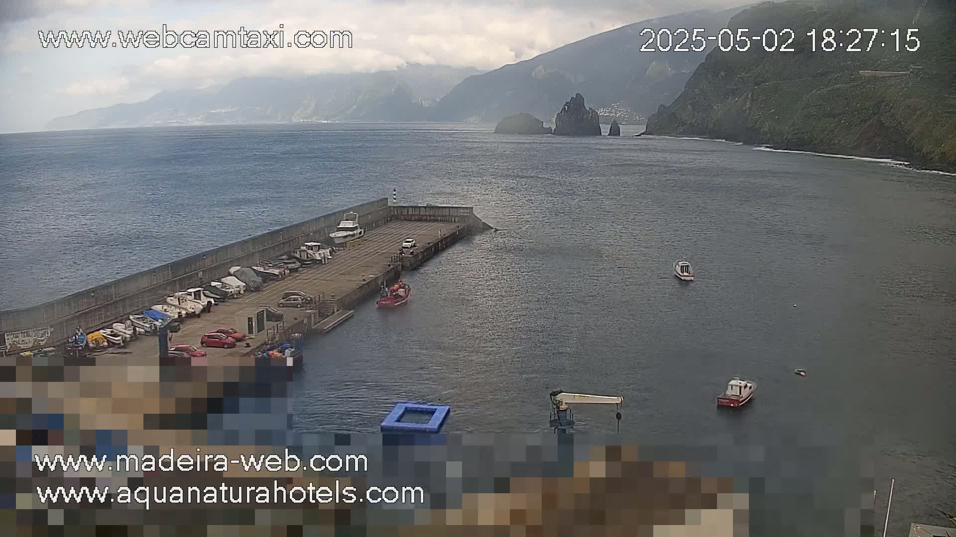 A harbor with several boats docked, a few more in the water, and a mountainous coastline under a cloudy sky.