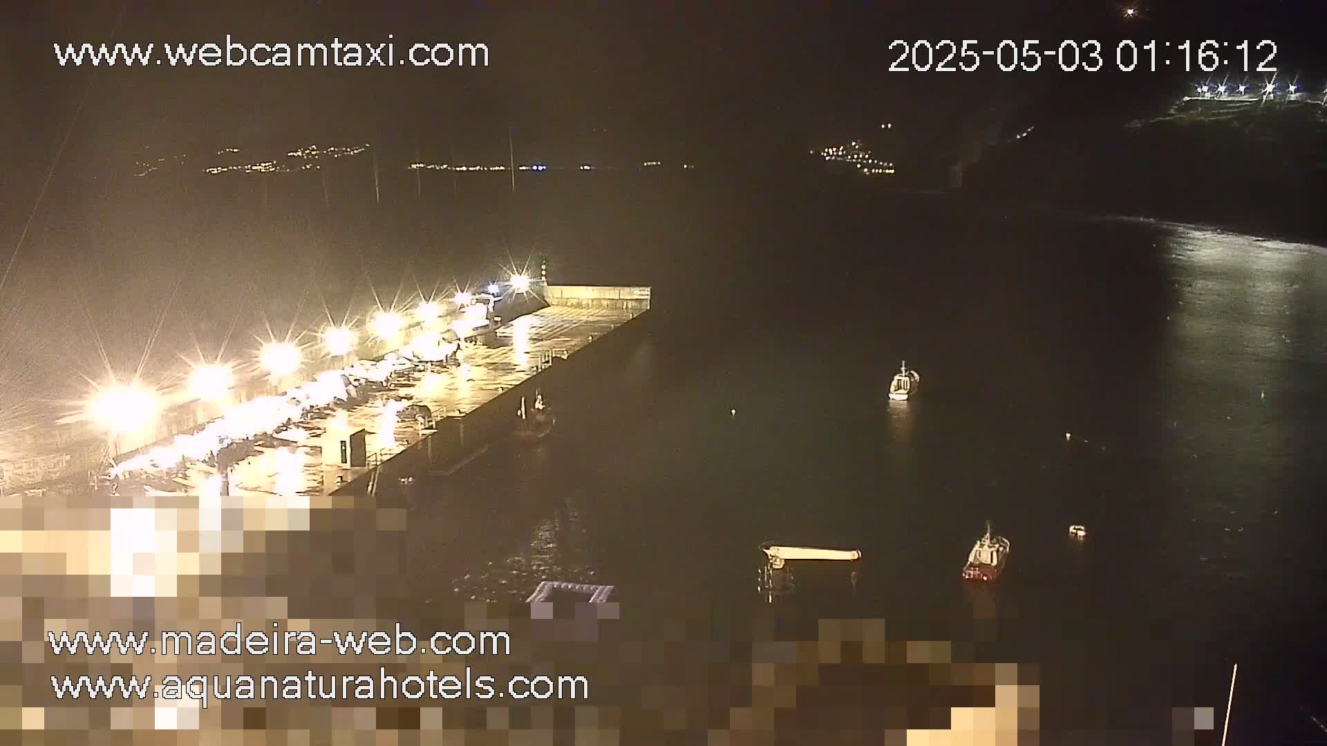 A brightly lit harbor at night shows several boats moored at a pier, with a few more boats and a calm, dark body of water visible beyond.