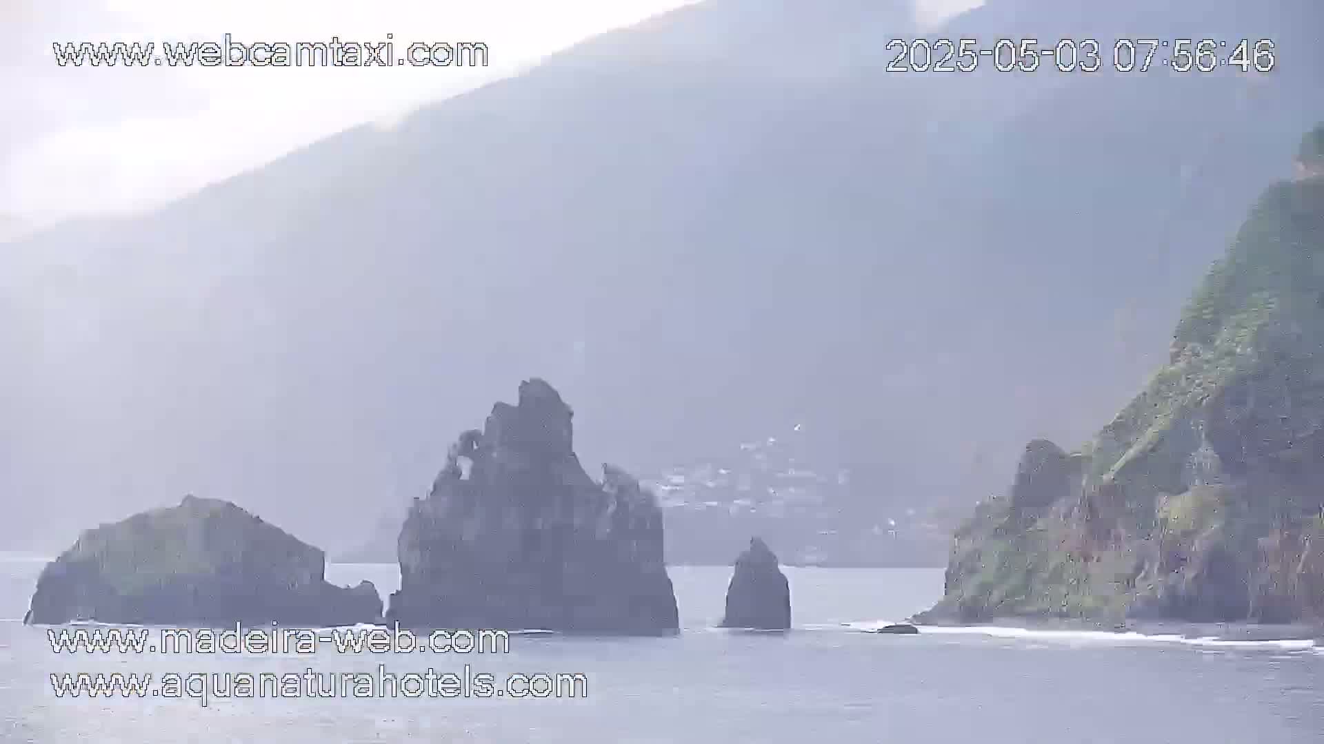 A hazy view shows dark, rocky formations jutting out of a body of water near a cliffside.