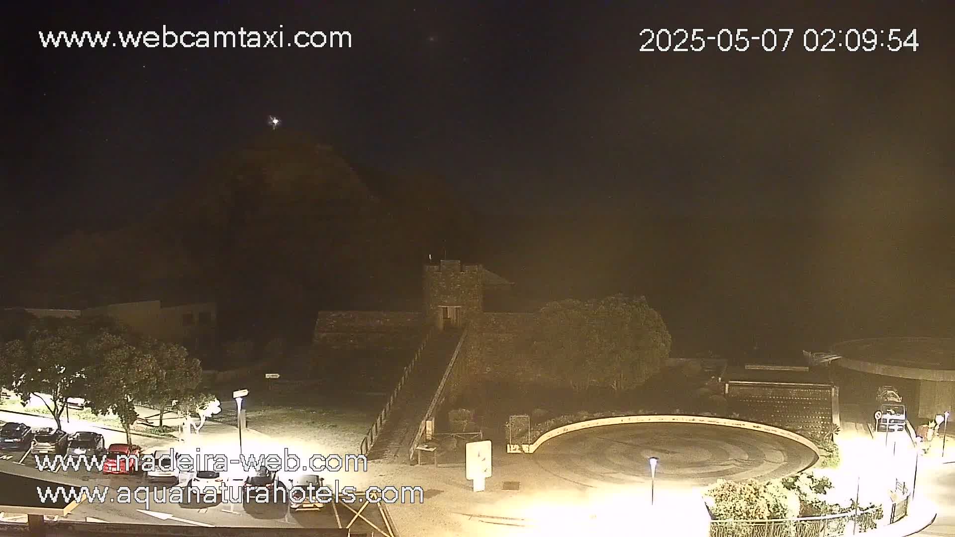 A nighttime view of a stone structure, possibly a castle or fort, situated on a hill overlooking a circular roadway and parking lot under a clear, dark sky.