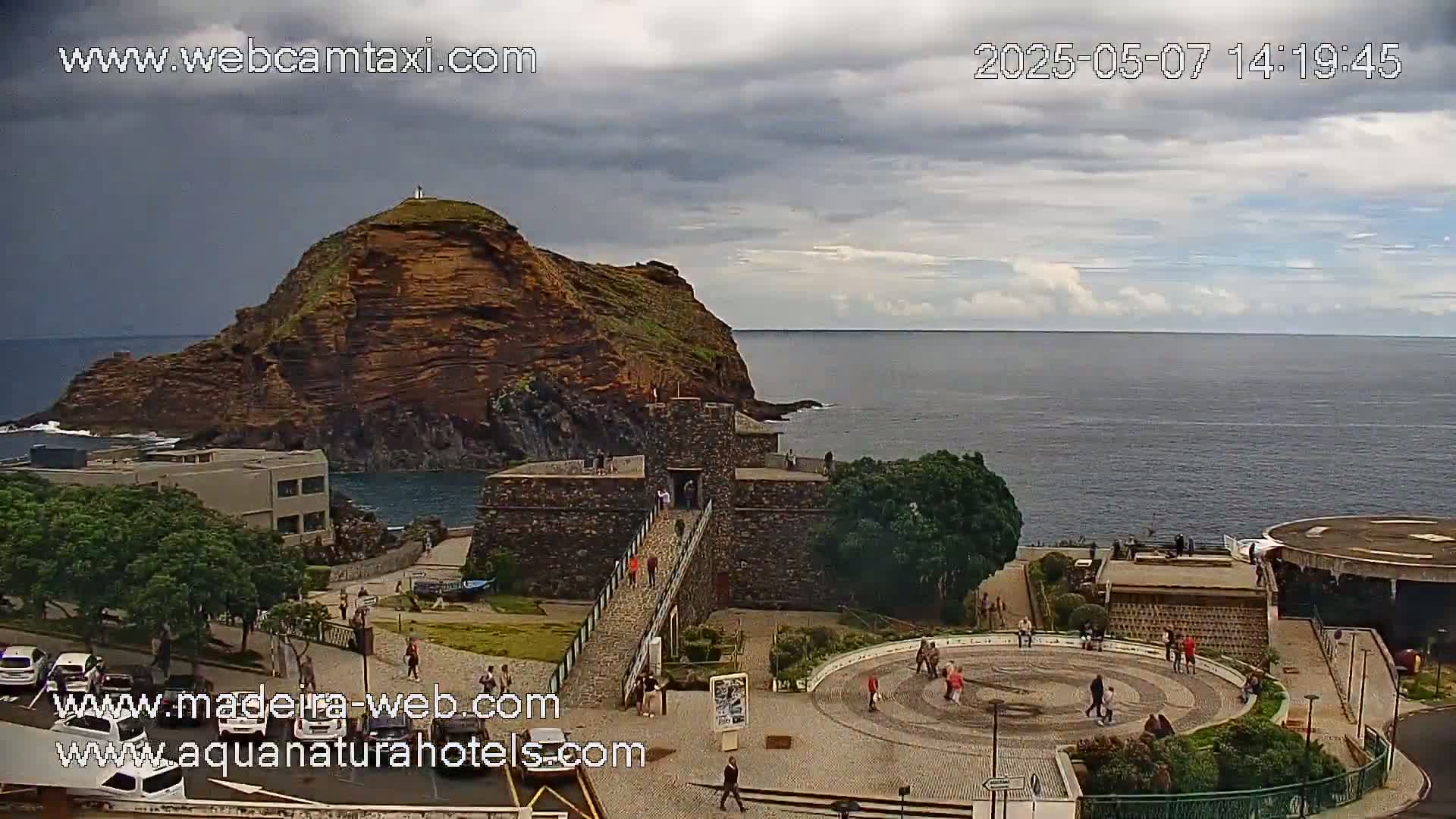A stone fortress with a bridge connects to a rocky hill overlooking a calm ocean under a partly cloudy sky.