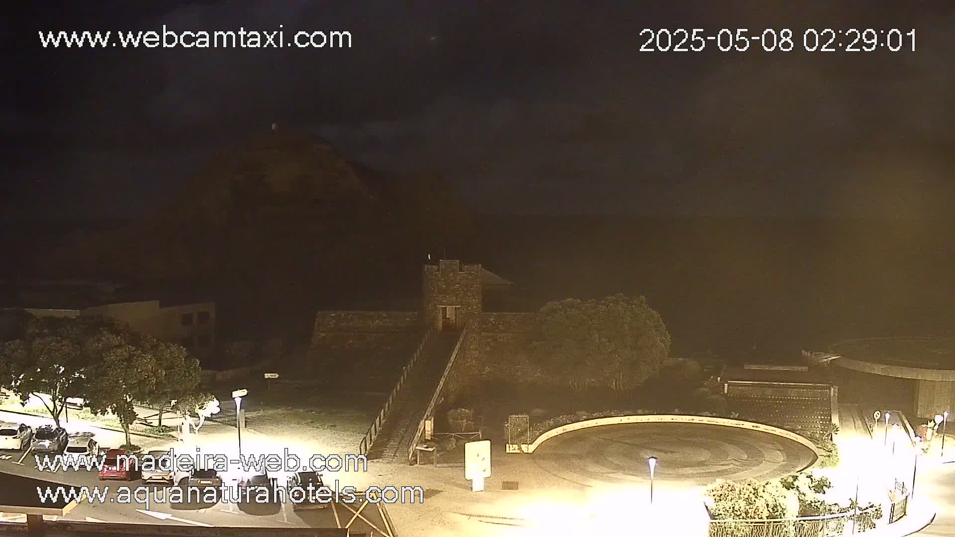 A nighttime view of a stone structure near a body of water under a cloudy sky, with dimly lit roads and vegetation surrounding it.