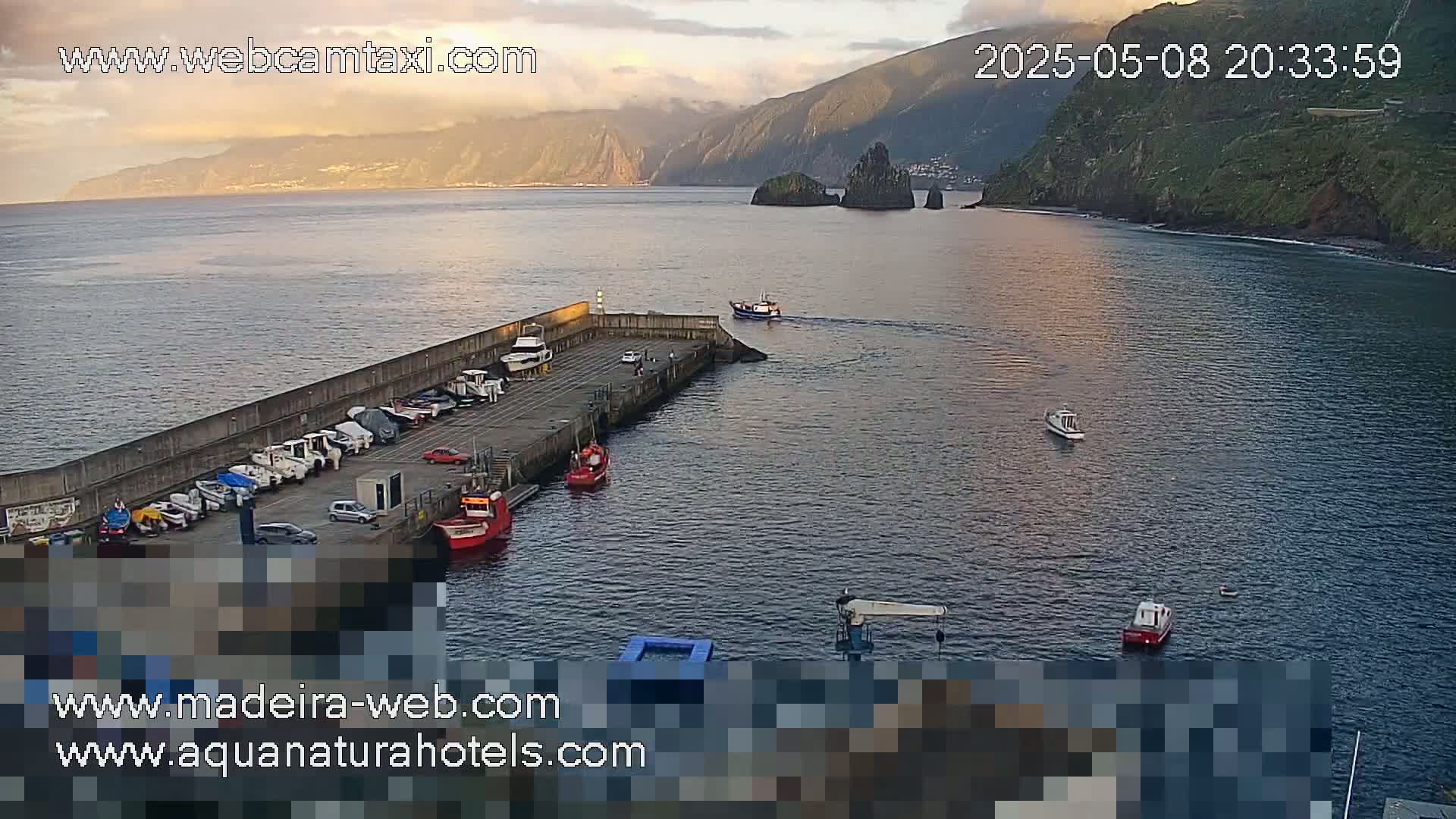 A calm harbor with several small boats docked and a few others in the water, situated next to a rocky cliff face under a partly cloudy sunset sky.