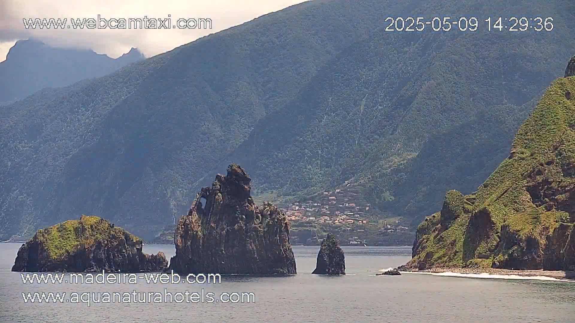 A coastal view shows dark rocky formations, including several prominent stacks, near a green, mountainous shoreline under a mostly clear sky.