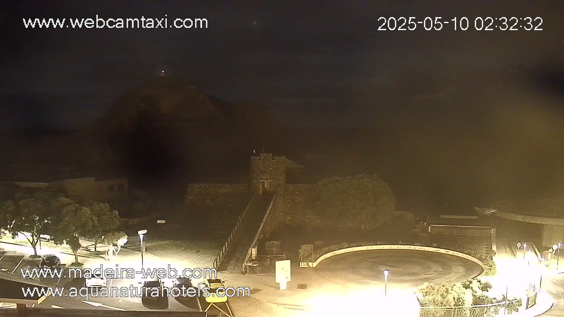 A nighttime view shows a stone structure on a hilltop, a circular plaza with cars, and trees under hazy conditions.