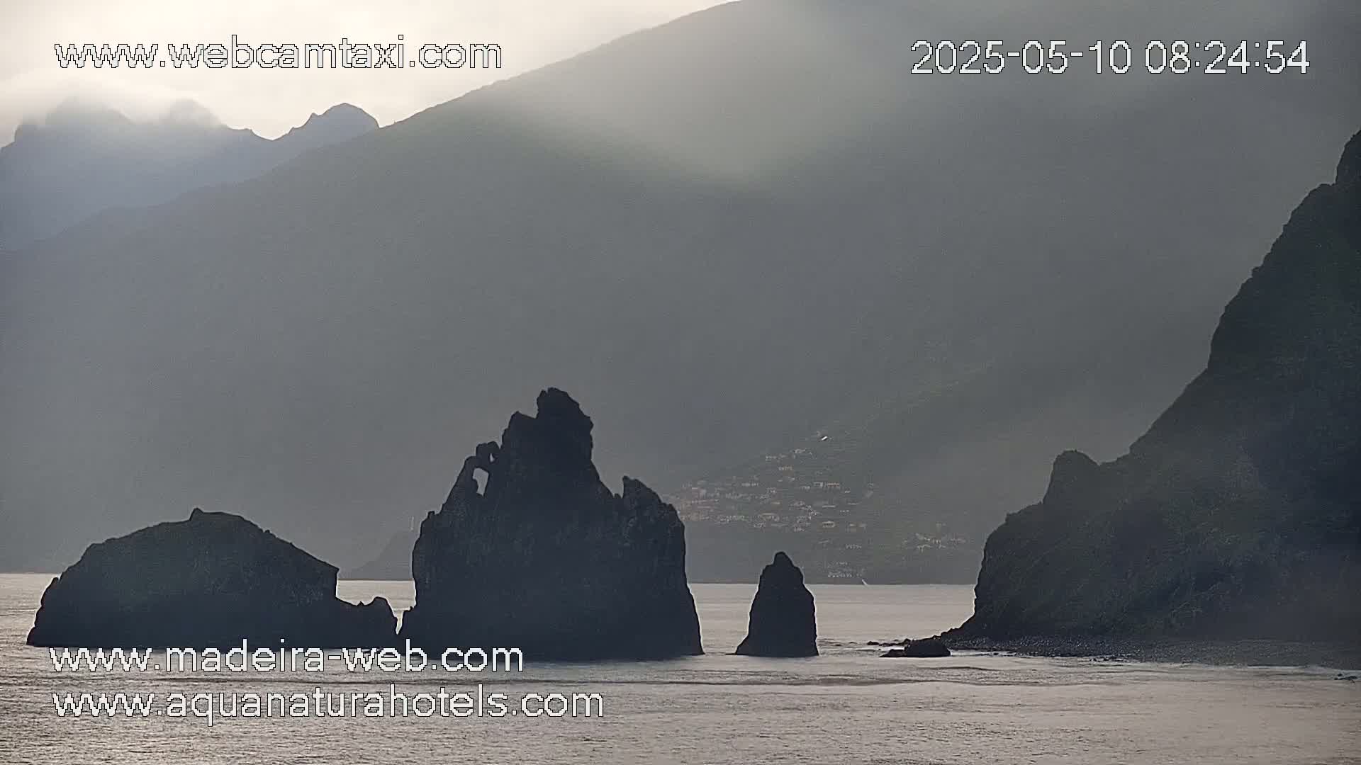 Dark, rocky formations rise from a calm sea under a hazy, overcast sky.