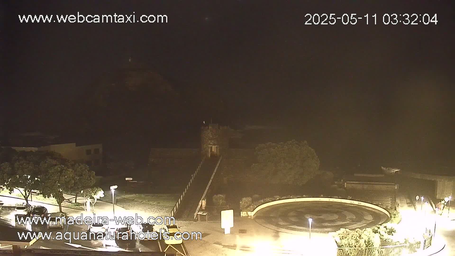 A nighttime, dimly lit outdoor scene shows a stone structure with a ramp, surrounded by landscaping and a circular plaza.
