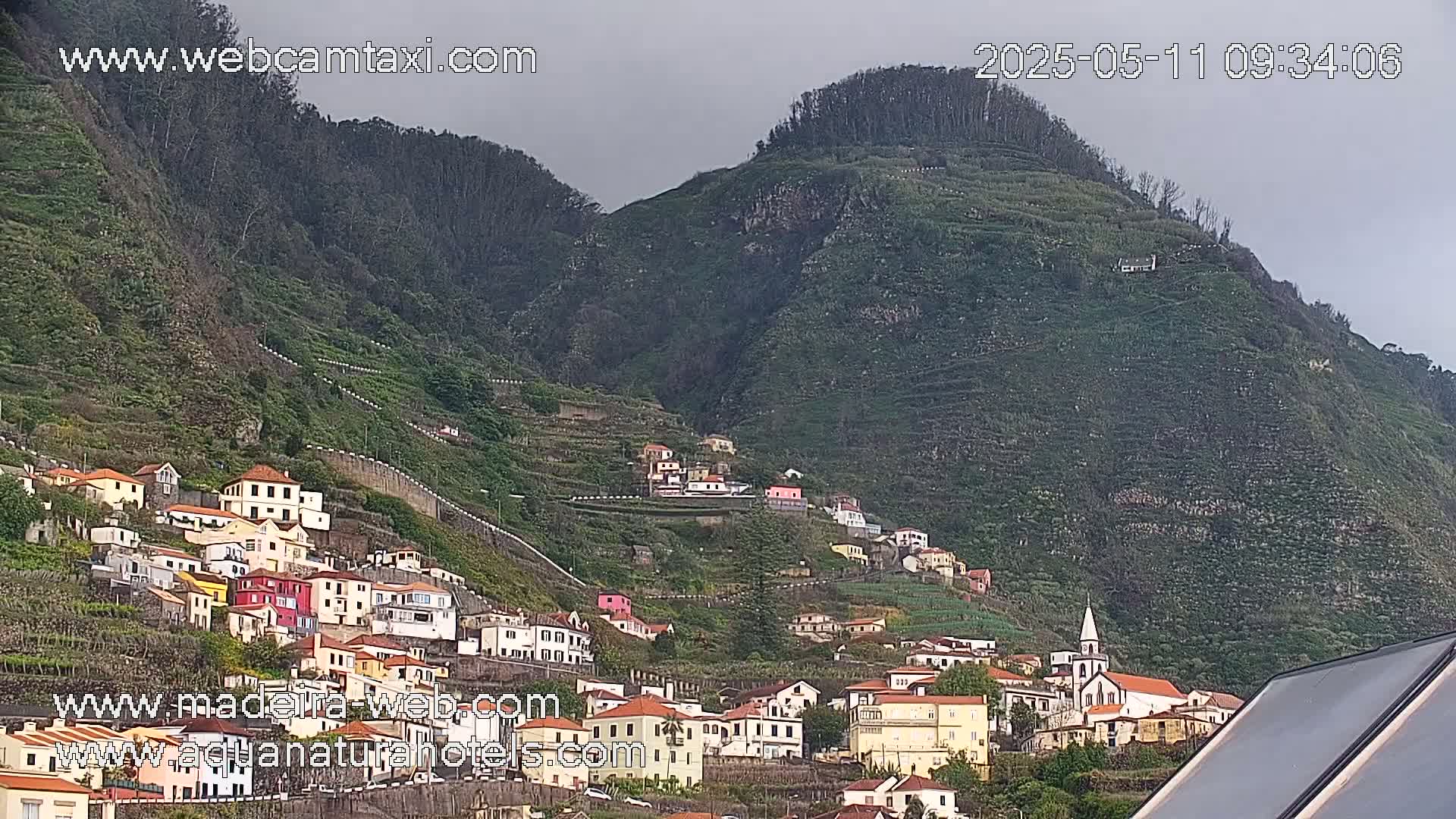 A hillside village of colorful houses clings to a steep, green mountainside under an overcast sky.