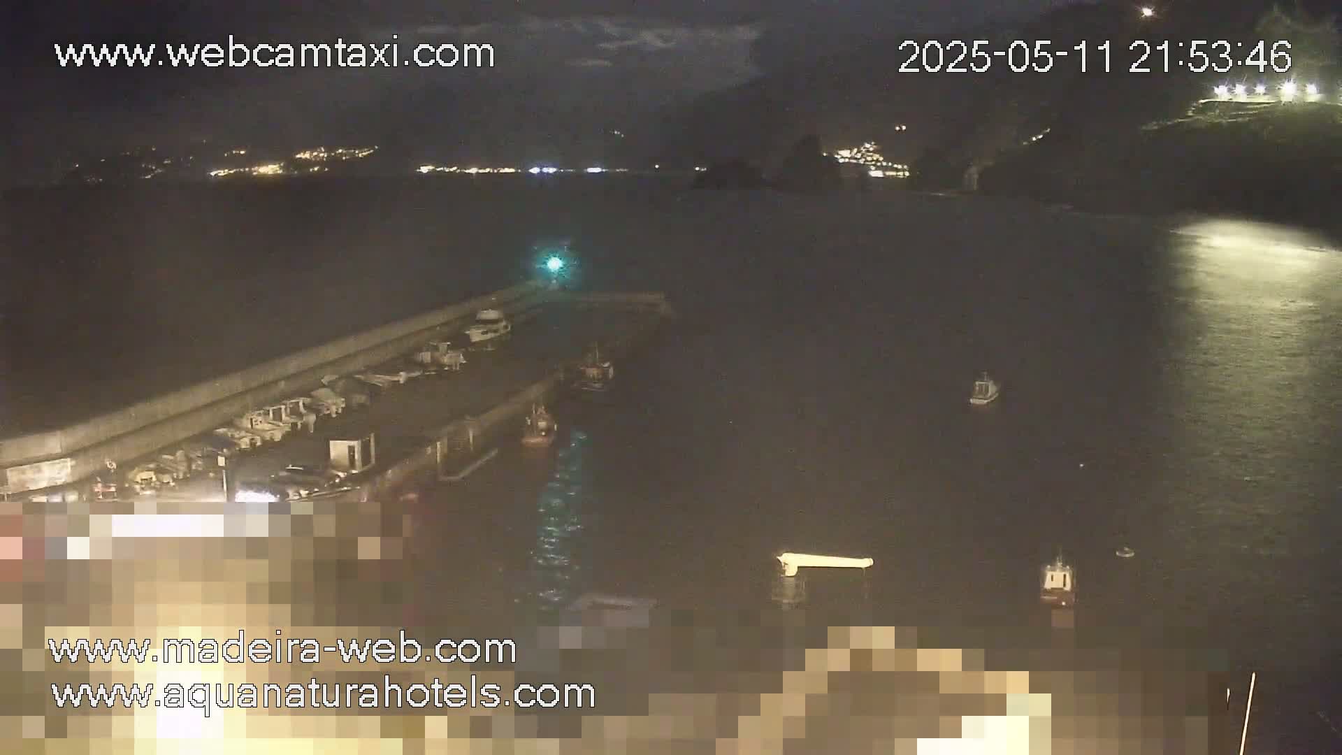 A nighttime view of a harbor with several boats moored, illuminated by lights from the shore and a dark body of water extending to a distant, faintly lit coastline.