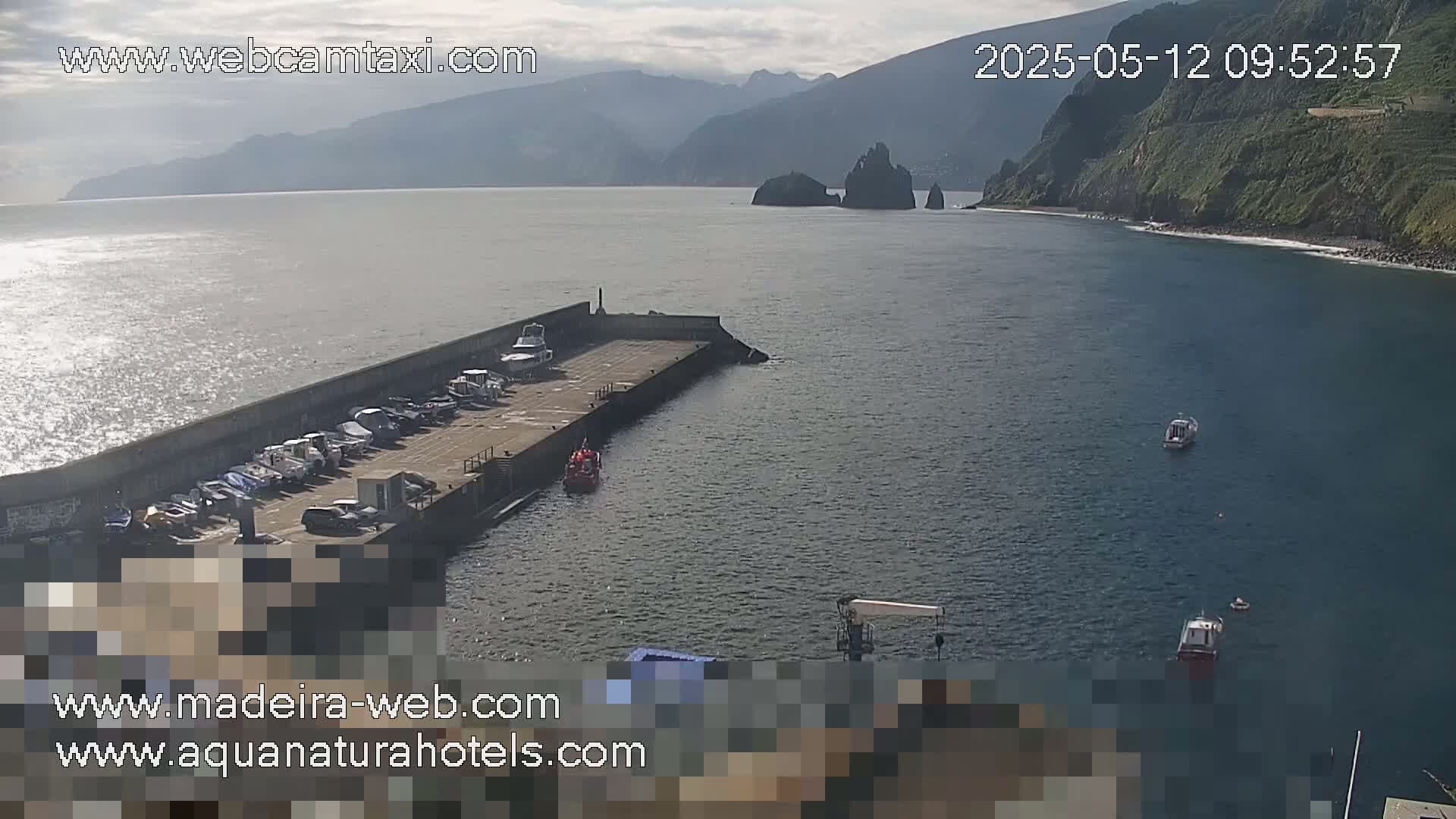 A mostly sunny day shows a small harbor filled with boats, next to a rocky coastline and mountainous landscape.