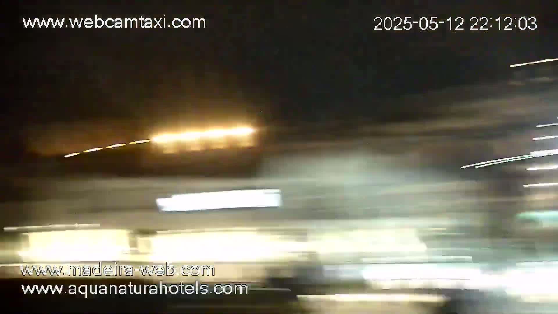 A blurry nighttime image shows streaks of light from moving vehicles, possibly on a highway, with some illuminated buildings in the background.