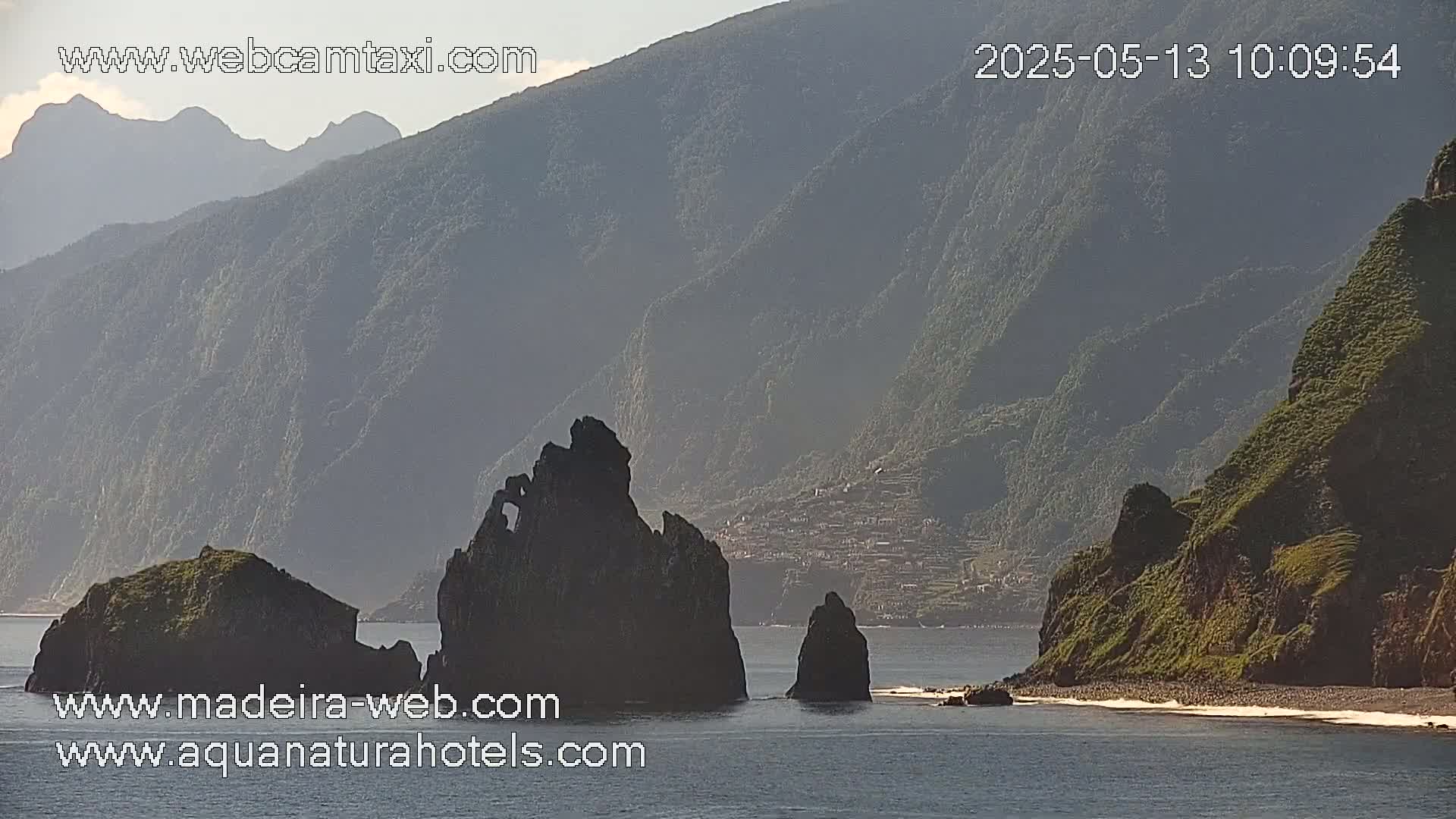 A hazy, sunny day reveals a rocky coastline with several prominent dark rock formations in the foreground, and a mountainous backdrop with a small village visible in the middle distance.