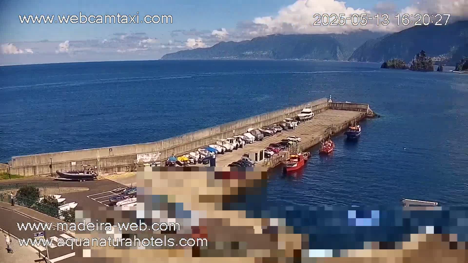 A mostly sunny day shows a harbor with numerous boats and cars parked along a concrete wall, overlooking a calm ocean and distant mountains.