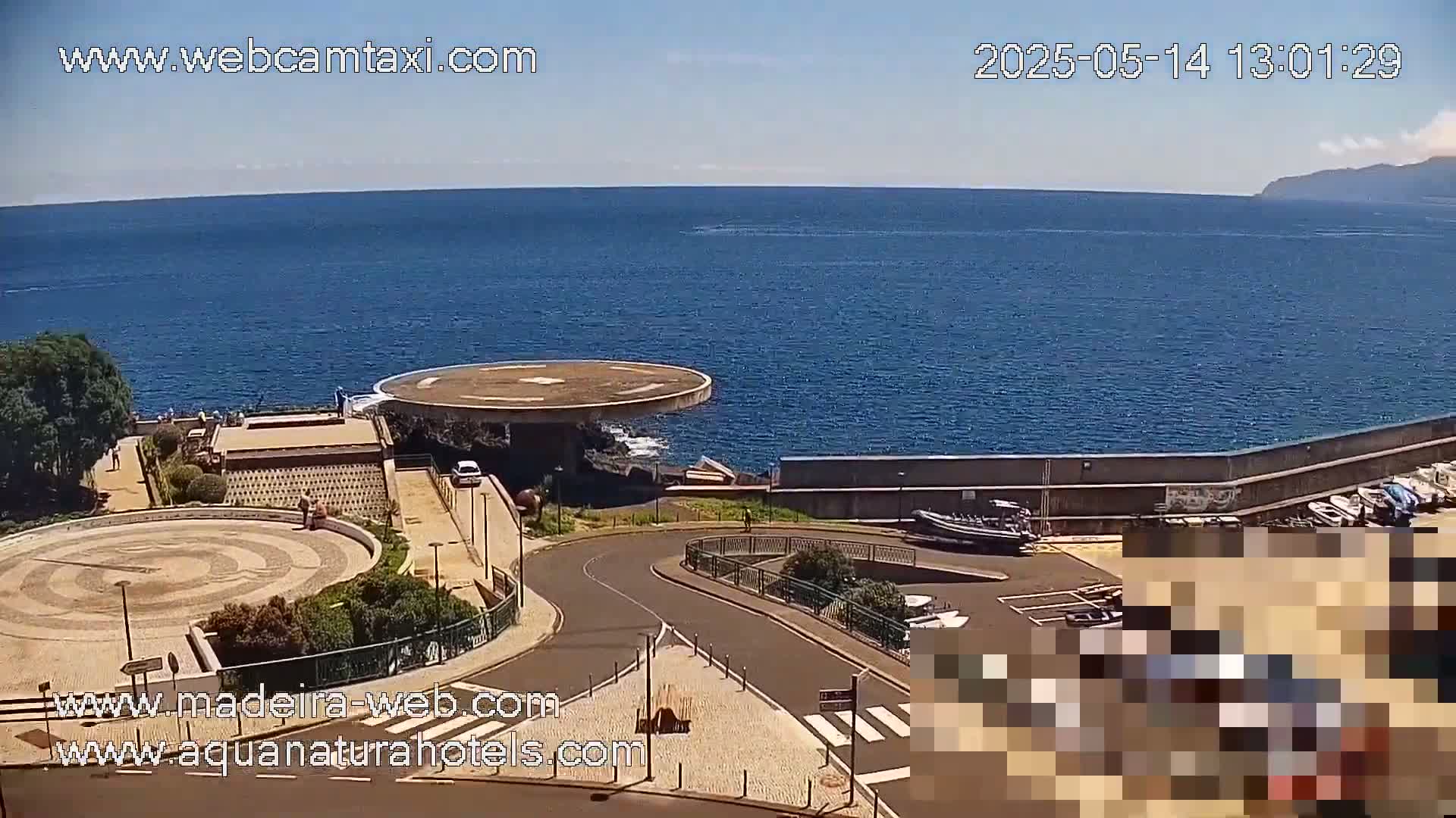 A coastal overlook on a sunny day shows a circular plaza, a helipad, a roadway, a parking lot with boats, and a calm ocean stretching to a distant mountain.
