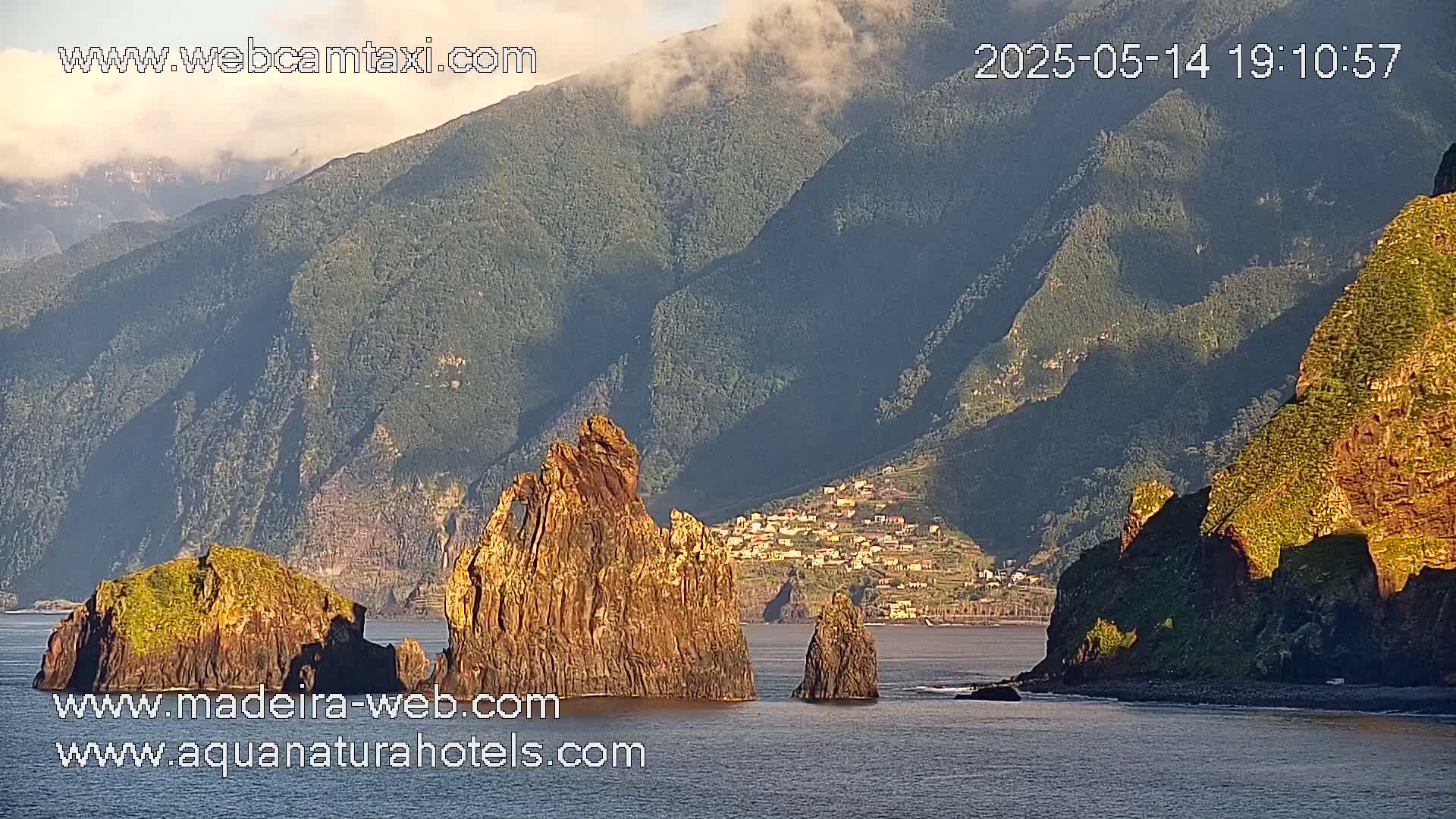 A tranquil coastal scene shows several rocky outcrops in calm water at the foot of a sunlit mountain range under partly cloudy skies.
