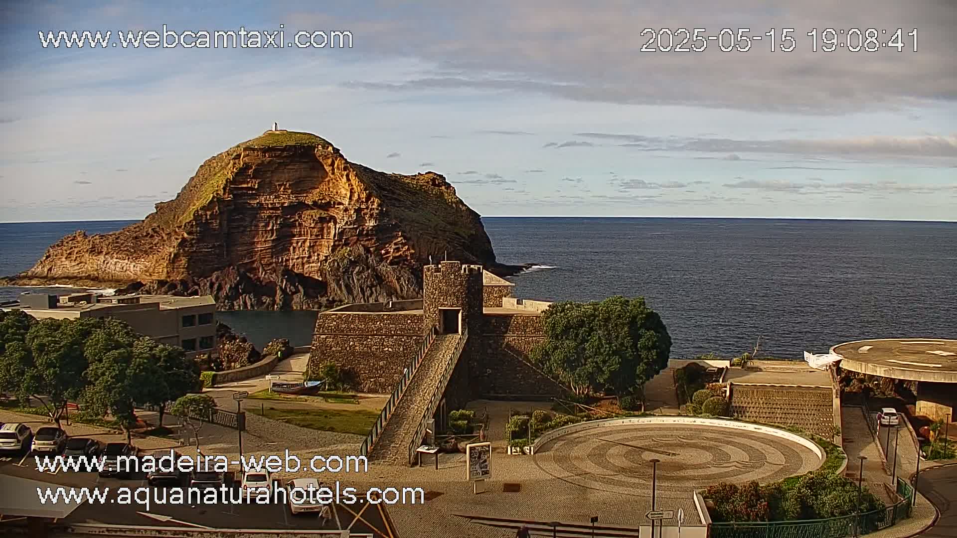 A stone structure and a rocky island jut out into a calm ocean under a partly cloudy sky.