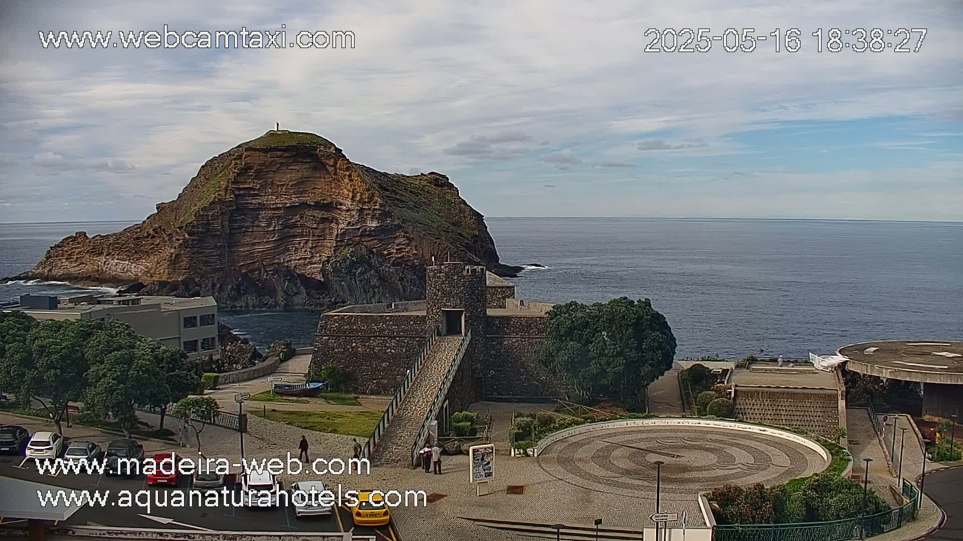 A coastal scene under partly cloudy skies shows a rocky, reddish-brown island connected to the mainland by a stone structure, with a circular plaza and parking lot visible in the foreground.