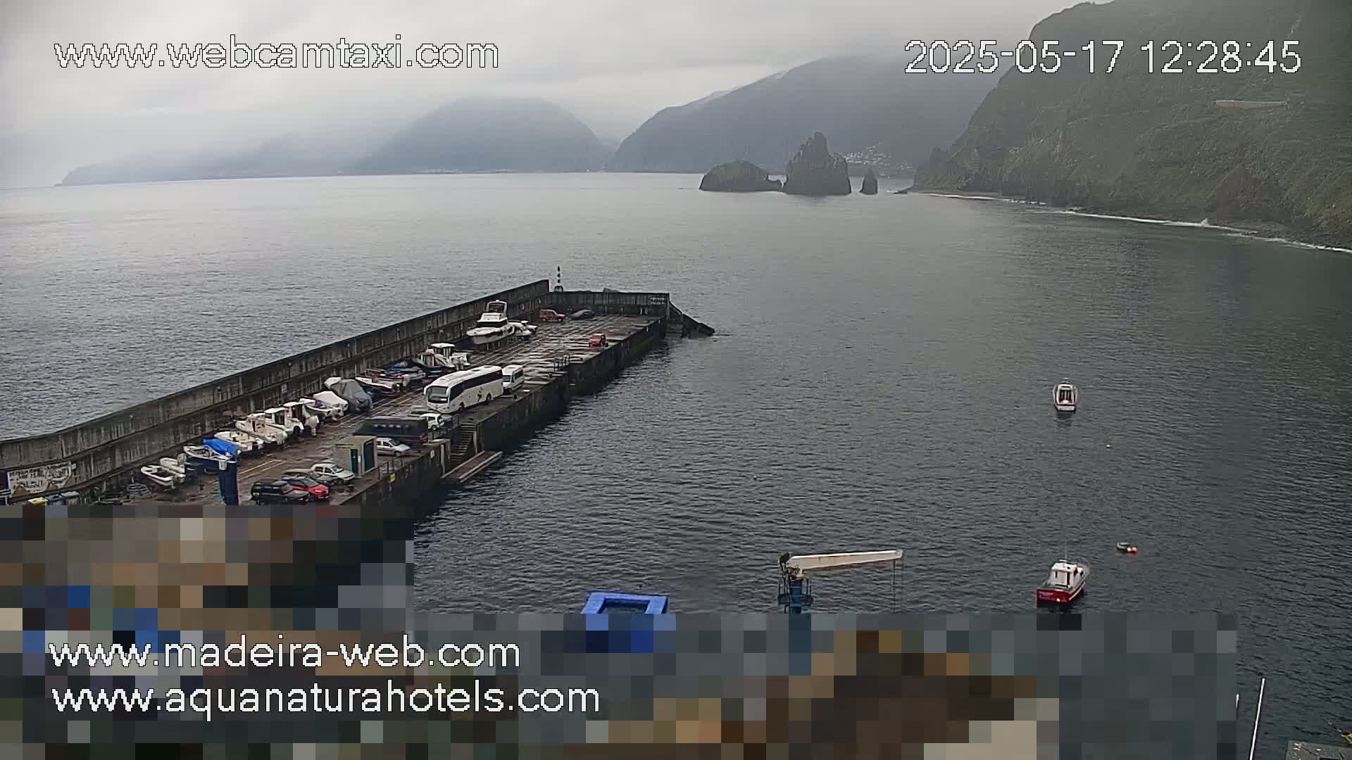 A harbor filled with several boats and cars under an overcast sky, with mountainous terrain visible in the background.