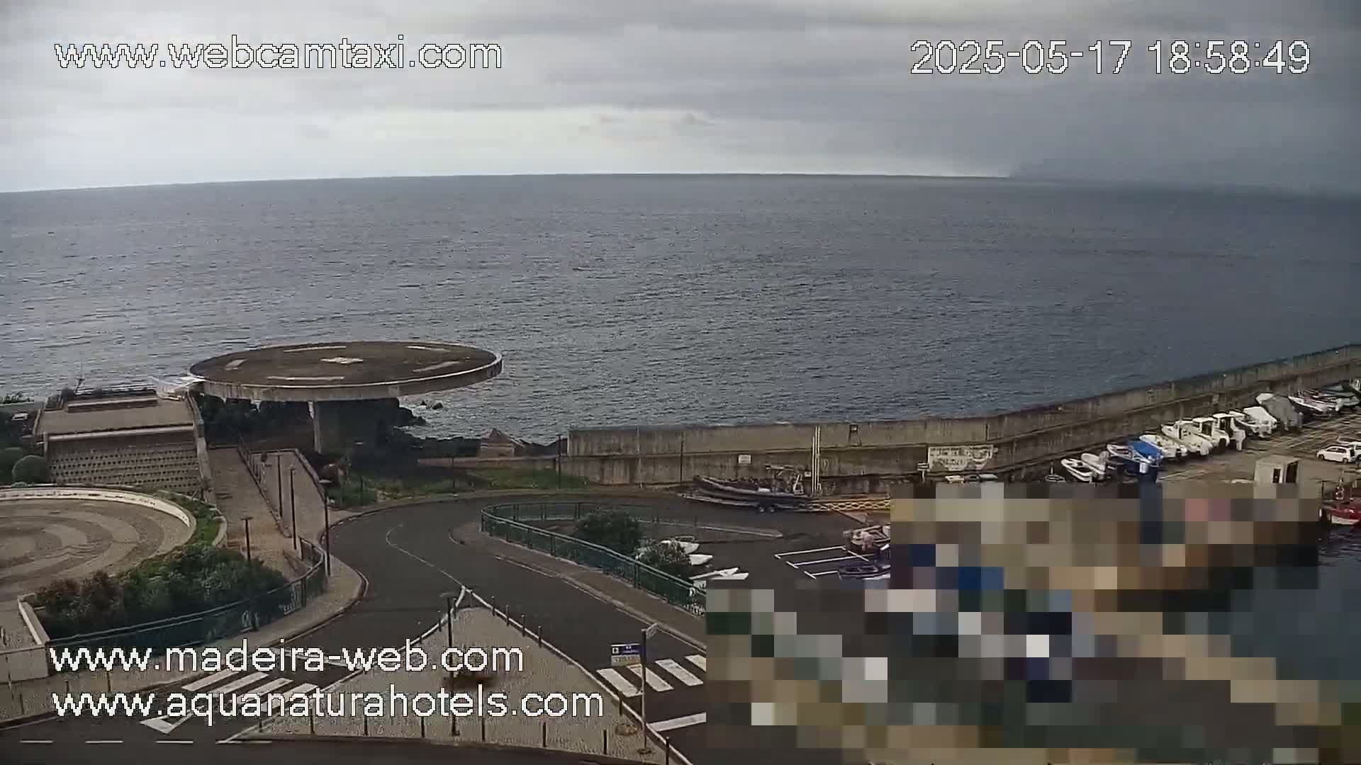 A coastal scene under an overcast sky shows a helipad, a harbor with several boats, and a road curving around a seawall overlooking a calm ocean.