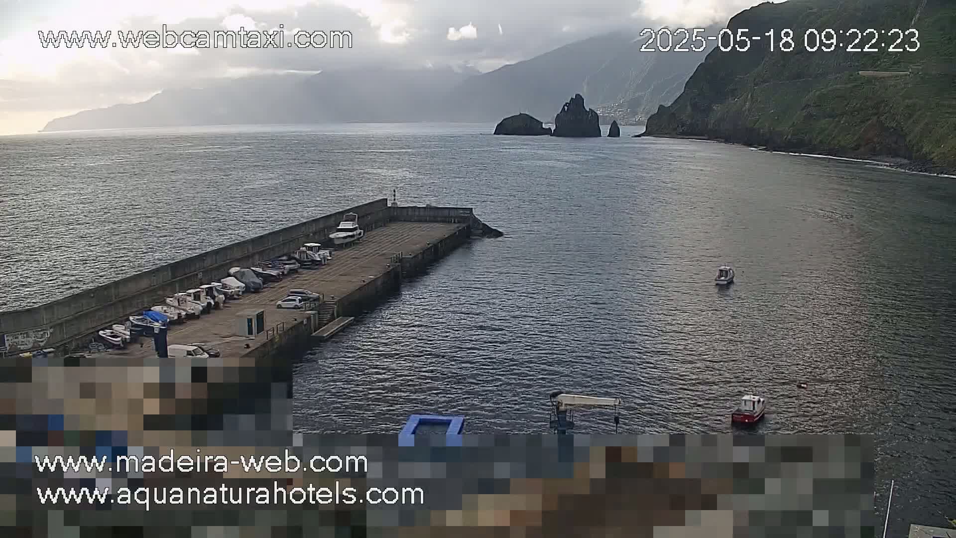 A harbor with several boats docked, situated against a mountainous coastline under an overcast sky.