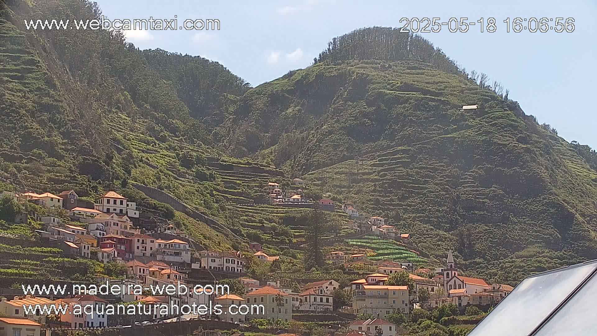 A hillside village with terraced farmland climbs a verdant mountain under a mostly sunny sky.